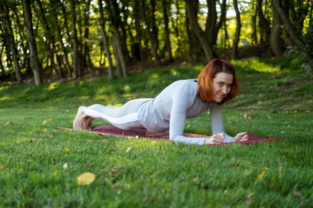 Millennial slim ginger woman doing yoga outdoor on nature in city in autumn sunny day on sunrise.