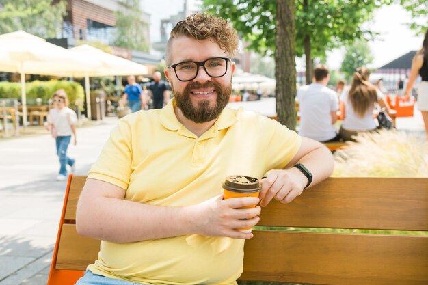 Foto millennial sorridente studente uomo in maglietta gialla polo occhiali seduto su una panchina bere caffè fare colazione riposare rilassarsi al sole primavera verde parco cittadino all'aperto sulla natura concetto di svago urbano