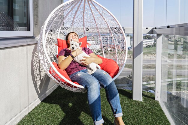 Millennial woman with white puppy sitting in modern hanging chair on balcony of apartment building and smile