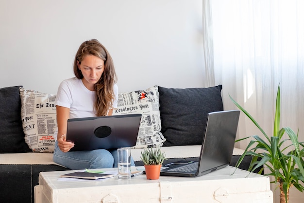 Millennial woman works at home,  seats close to Laptop computer with notebook, glass of water on white table in home workspace.