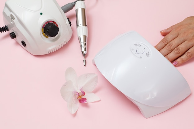 Milling cutter, led UV lamp and a female hand on a pink background. A set of cosmetic tools for professional hardware manicure. Top view