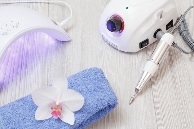 Milling cutter, turned on led UV lamp and a towel with orchid on gray wooden background. A set of cosmetic tools for professional hardware manicure. Top view