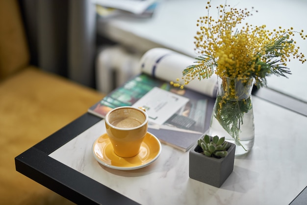 花瓶のミモザとカフェやレストランのテーブルの上の黄色のカップのコーヒー