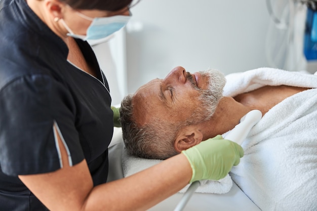 Mindful female beautician wearing a face mask while conducting skin rejuvenation procedure for an elderly person