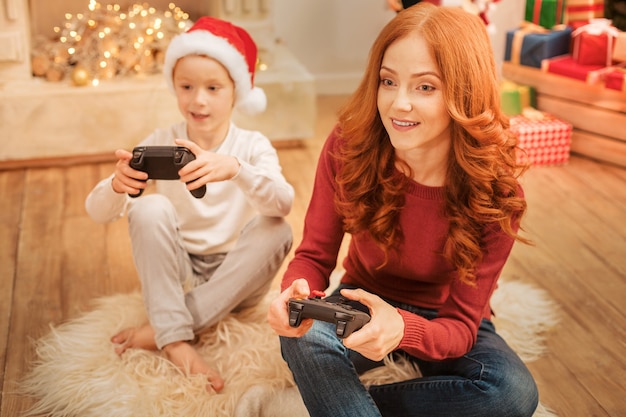 mindful mature lady sitting on the carpet and looking at a TV screen with excitement while having fun and video gaming with her child.