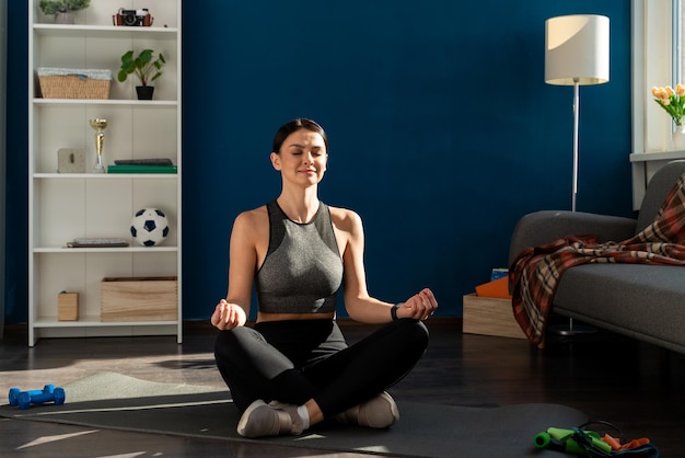 Mindful meisje zit met gekruiste benen bij de bank, beoefent yoga en geniet van meditatie. Jonge vrouw met gekruiste benen en gesloten ogen die thuis mediteren