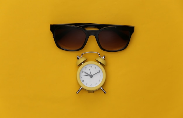 노란색 배경에 미니 알람 시계와 선글라스. 여름 휴가 시간입니다.