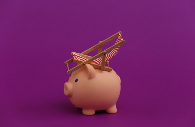 Mini beach deck chair and piggy bank on purple. Symbol of beach holidays and resort.