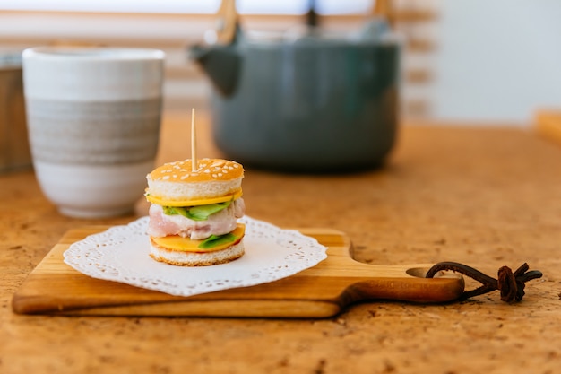 Mini Chicken Burger on wooden chopping board with blur tea cup and mug in the background.