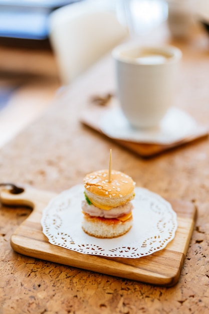 Mini chicken burger sul tagliere di legno con sfocatura tazza di tè e tazza in background.