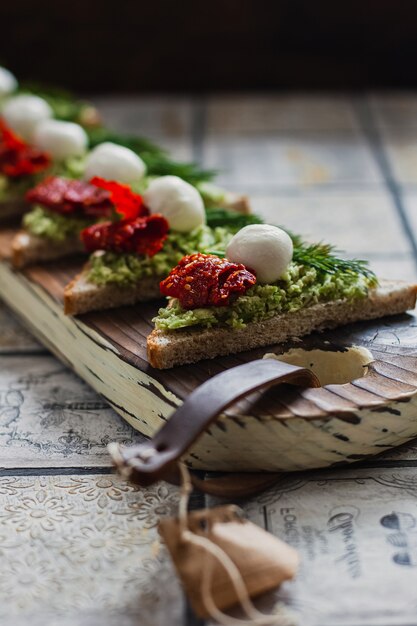 Mini driehoek canapés bruschetta met avocado, mozarella en gedroogde tomaten op het rustieke bord op de vintage betegelde achtergrond.