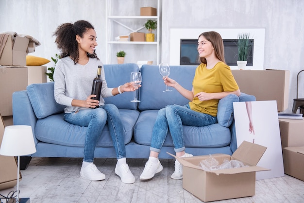 Mini-feest. Vrolijke jonge meisjes die op de bank zitten en op het punt staan wat wijn te drinken terwijl ze vieren dat ze naar een nieuw appartement gaan