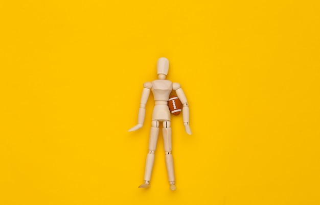 Mini houten pop met rugbybal op een gele achtergrond