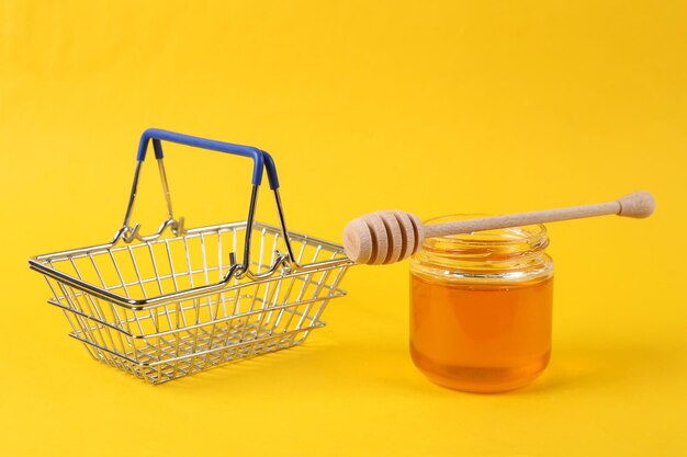 Mini shopping basket with bee honey jar and honey wooden spoon on yellow background