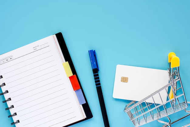 Мини-корзина или тележка с пустой кредитной картой, спиральной тетрадью и ручкой на синем фоне