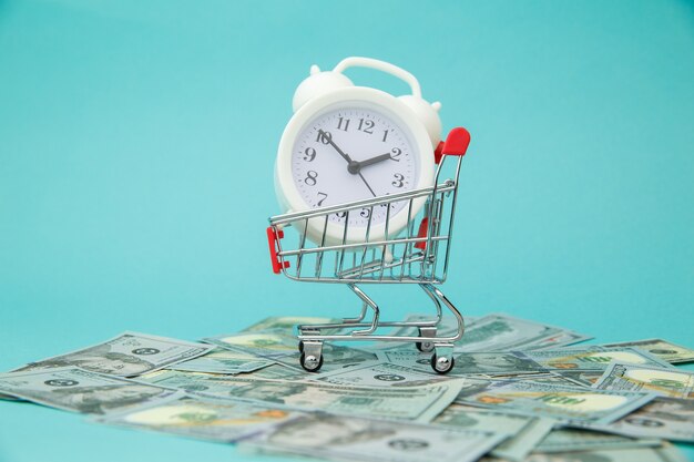 Mini shopping cart with white alarm clock on blue.