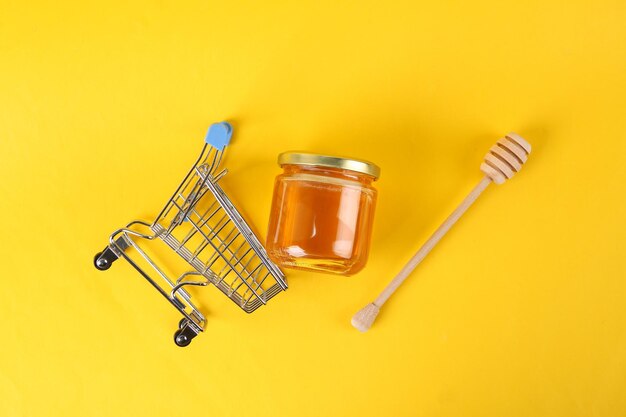 Мини-тележка для покупок и банка пчелиного меда с медовой деревянной ложкой на желтом фоне