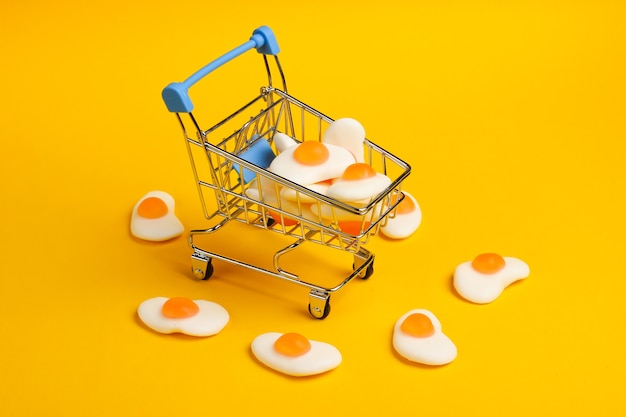 Mini shopping trolley with marmalade fried eggs on a yellow background. Sweets Shopping, Pastel Color Trend, Food Concept