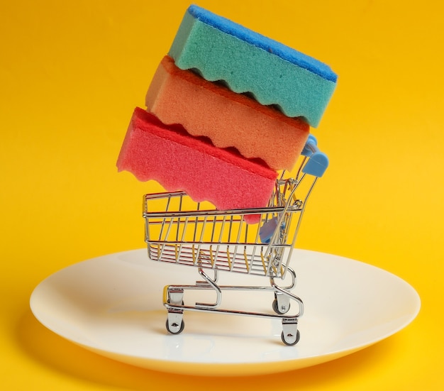 Mini shopping trolley with sponges for washing dishes on a plate. Yellow background