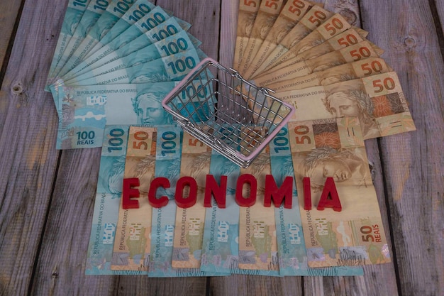 Mini supermarket basket on banknotes of 50 and 100 reais on a wooden background written in Portuguese Economia