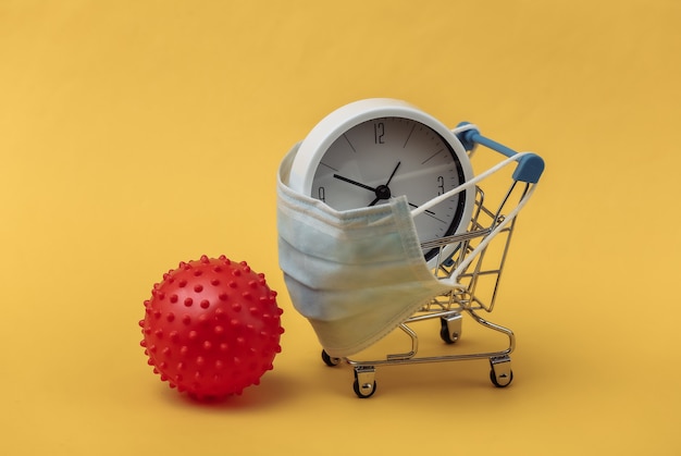사진 의료용 마스크에 시계가 달린 미니 슈퍼마켓 트롤리, 노란색 배경에 바이러스 변형 모델.