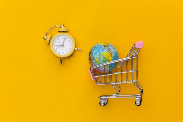 Mini supermarket trolley with globe and retro alarm clock on yellow background