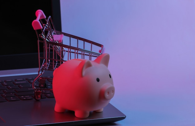 Mini supermarket trolley with piggy bank on laptop keyboard.\
neon gradient red-blue, ultraviolet light. online shopping