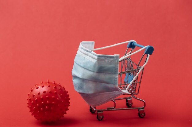 Mini carrelli per supermercati con maschera medica, modello di ceppo virale su sfondo rosso. fare la spesa durante la pandemia di covid-19