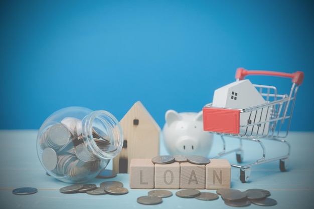 ミニ木造住宅モデルがショッピング カートに入っており、青い机の上の木のブロックにローンというテキストが表示されます。将来の銀行財務と会計のための不動産取引のアイデアの貯蓄です。