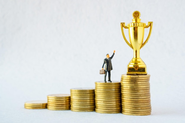 Miniature businessman and gold trophy standing on growing coins on white background The concept of competition and victory