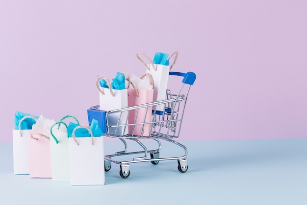 Miniature cart filled with paper shopping bags on blue surface