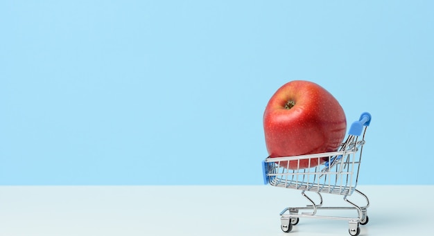 Miniature iron shopping cart with an apple on a blue background. Healthy food, food delivery, copy space