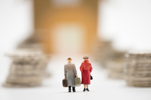 Behind miniature man and woman model hand carry suitcase and walk through coins stacking for move to new house. 