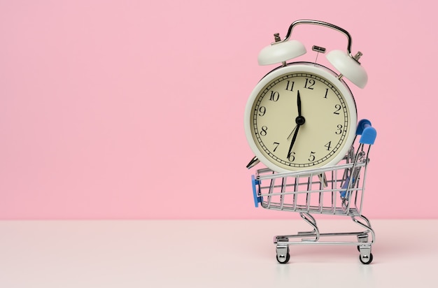 Miniature metal shopping cart on wheels and in the middle of a round alarm clock on a white table. Start of discounts, sale, copy space