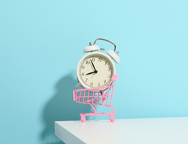 Miniature metal shopping cart on wheels and in the middle of a round alarm clock on a white table. Start of discounts, sale