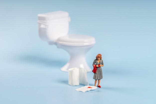Миниатюрная модель женщины стоит возле игрушечного туалета с туалетной бумагой с кровью