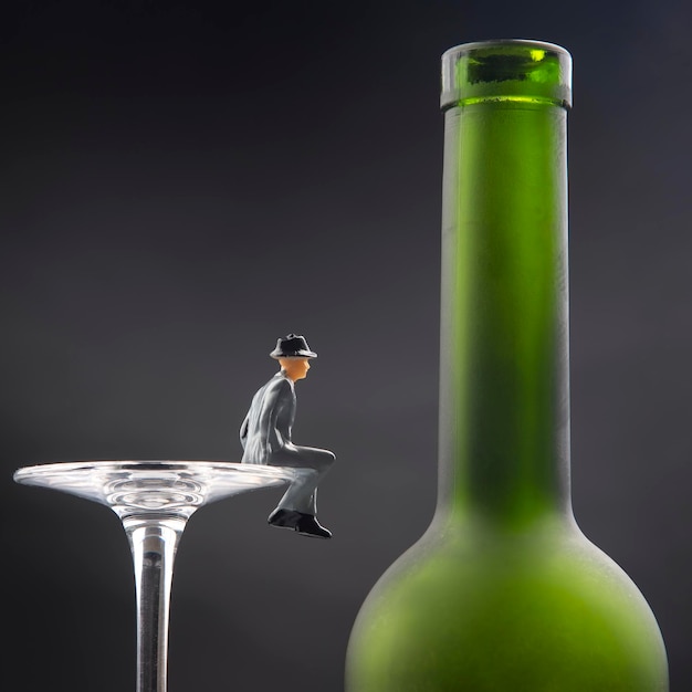 Foto persone in miniatura. concetto di problema di dipendenza da alcol. l'uomo alcolizzato si siede sul bordo di un bicchiere di vino vicino alla bottiglia