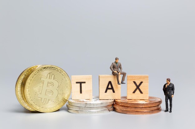 미니어처 사람들, 암호 화폐를 가진 사업가, 회색 배경에 나무 글자 TAX, Bitcoin 투자 및 세금 개념.