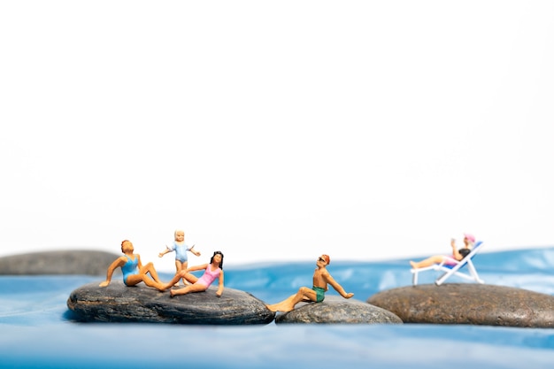 Miniature people, happy family sitting on the large rocks near the coast with blue sea and white background, Summer concept