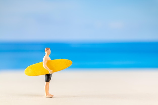 Foto uomo in miniatura in costume da bagno che tiene una tavola da surf gialla sulla spiaggia