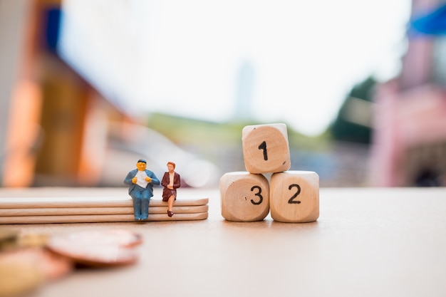 Miniature people, man and woman sitting with wooden number block and stack coins using as business a