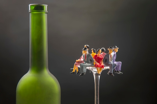 Miniature people sit on the edge of a wine glass near the bottle