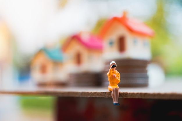 Miniature people, woman sitting in front of mini house on stack coins, using as business and propert
