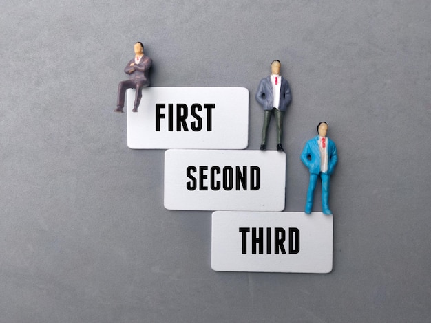 Miniature people and wooden board with word FIRST SECOND THIRD on a gray background