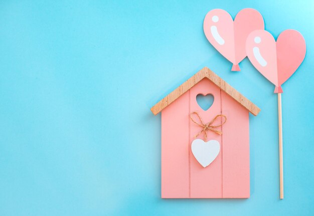 Miniature pink wooden flat toy house with hearts on blue background. Two heart-shaped balloons St. Valentine's Day composition.