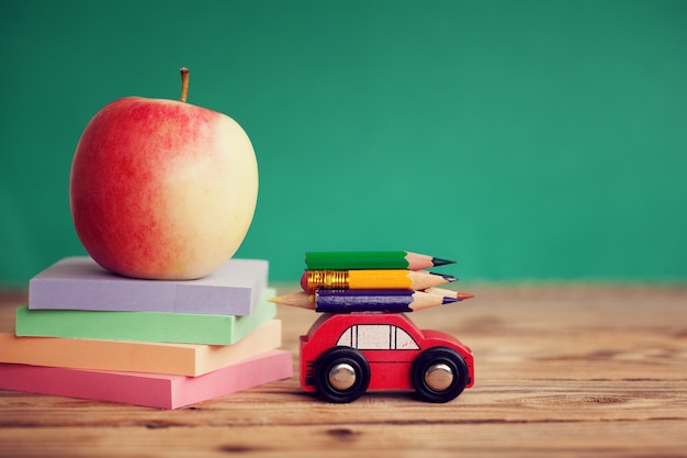 Automobile rossa miniatura che porta le matite variopinte e le carte rosse della pila e della mela sulla tavola di legno. torna al concetto di scuola