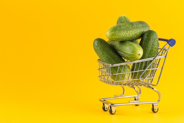 Miniature shopping cart filled with cucumbers on yellow background.