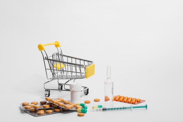 Miniature shopping cart with pills and medical syringe and ampoule on white background