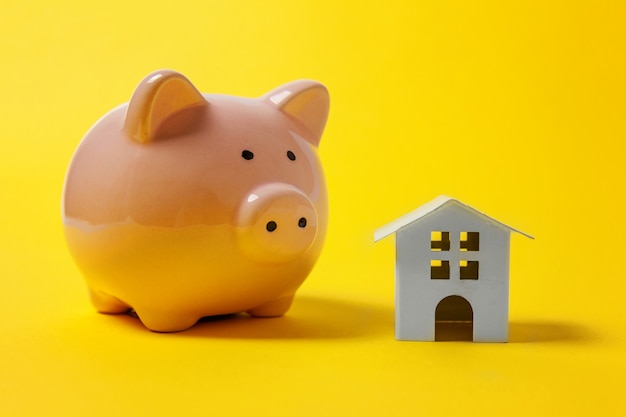 Miniature white toy house and piggy bank on yellow background