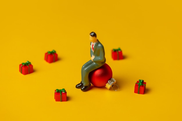 Miniatuurfiguur van man zit op kerstbal omringd door geschenken op geel oppervlak